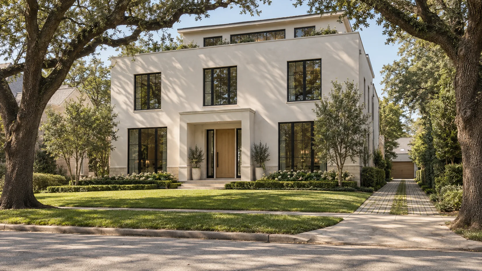 A two-and-a-half-storey house in cream plaster with matte black steel windows and a white oak front door under a plaster entry surround, a small top floor set back behind a parapet with a band of windows, seen across a street between two large live oaks, with a lawn and clipped hedges in front and a paver driveway with a grass strip running down the side to a garage set well back at the rear