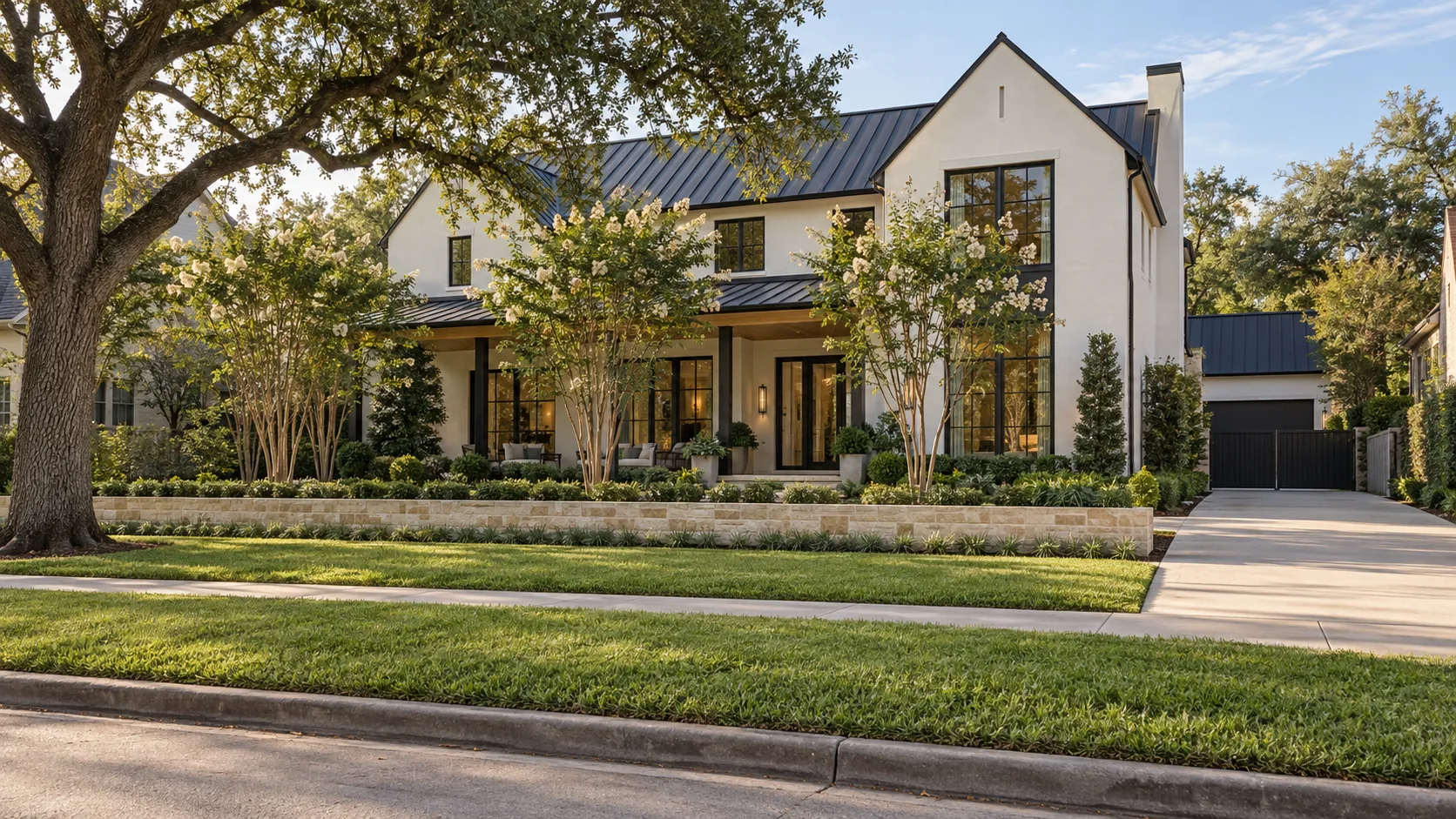Street frontage in morning light looking across a concrete curb and gutter, a wide grass verge and a concrete sidewalk to a low limestone planter wall, three crape myrtles in bloom and a two-storey white plaster house with a dark standing-seam metal roof, matte black windows and a covered porch, with a live oak at the left and a concrete driveway at the right
