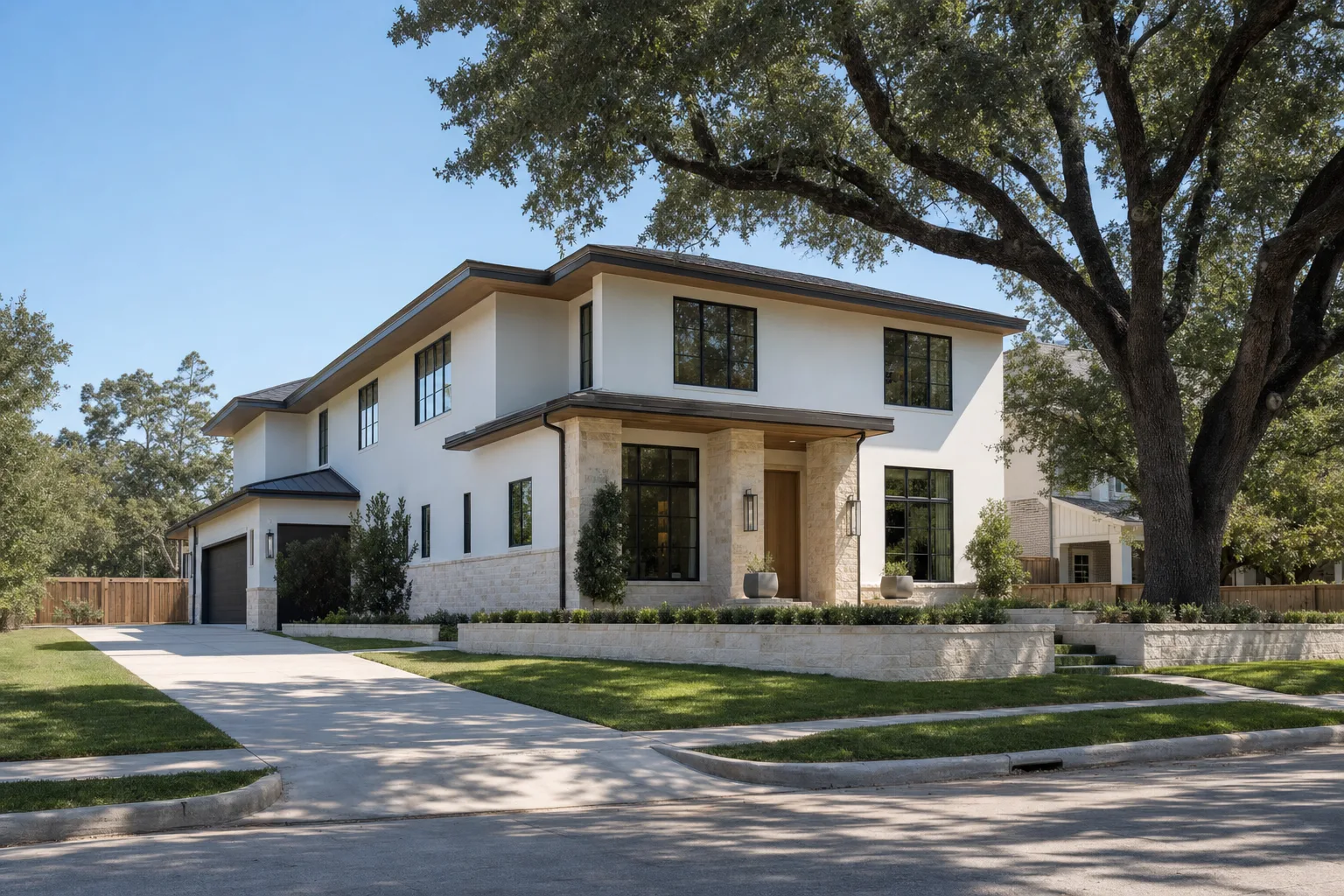 A two-storey modern house on a corner lot, white plaster above a limestone base with matte black windows, a low limestone garden wall along the front lawn, a concrete driveway running back to a side-facing garage, and a mature live oak overhanging the street