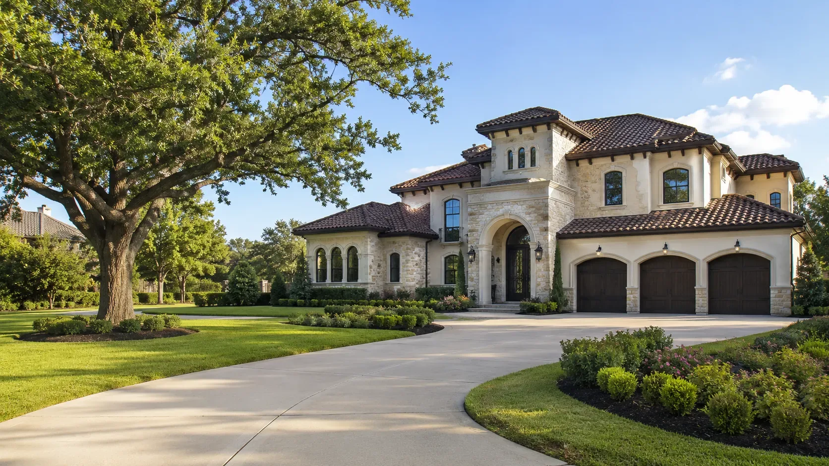 A large stone and stucco custom home in the Houston area with a tiled roof, arched entry and three-car garage, seen from a curving driveway beneath a mature oak on a clear day